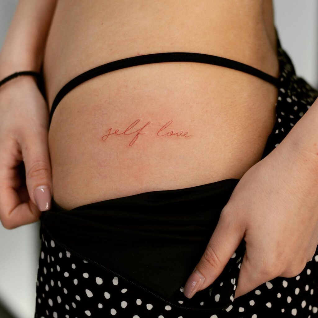 'Self Love' Text Script Lettering Tattoo on the Hip, Fineline Text in Striking Red: Self-Love Radiance 